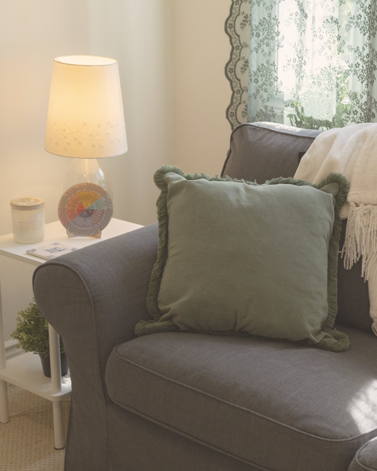 A cozy grey couch with a green pillow, a cream throw and a warm lamp by lace curtains in the Heart of Oak office in Oakville