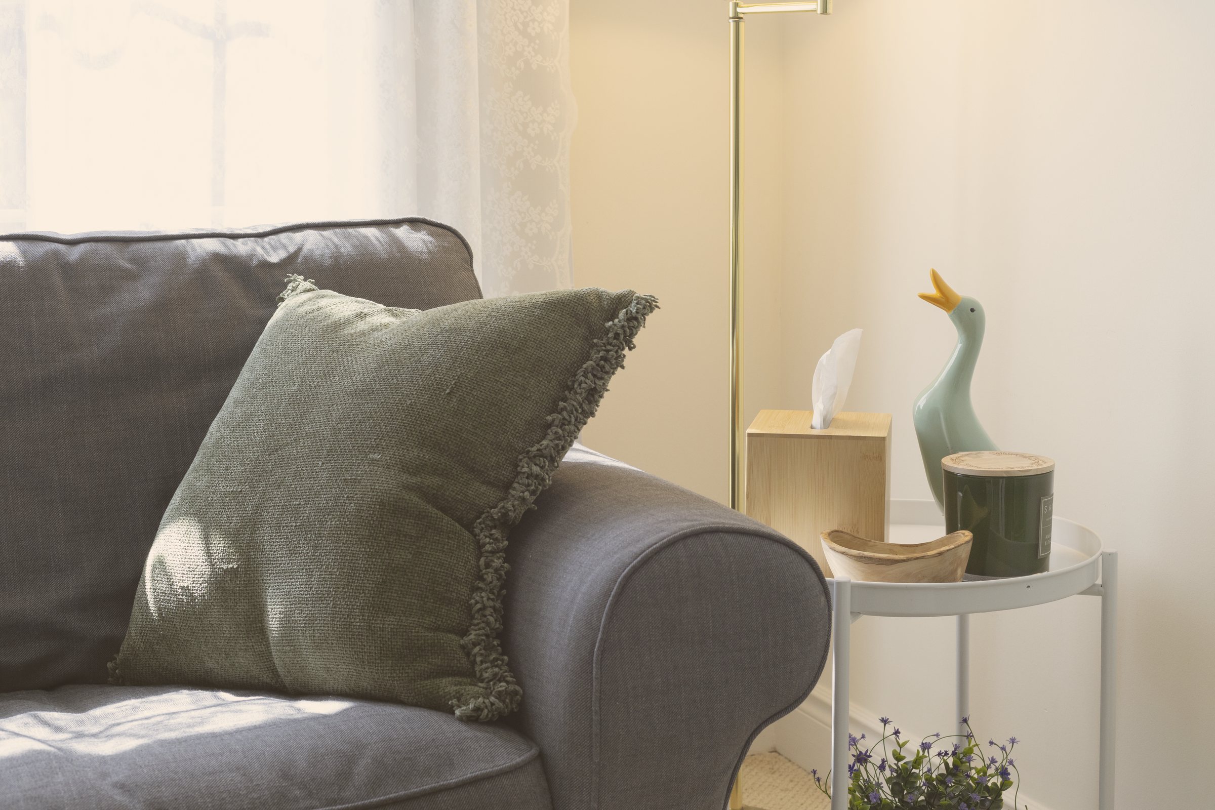 A grey couch with a green pillow beside a small side table with tissues, a candle and a ceramic duck in the Heart of Oak office