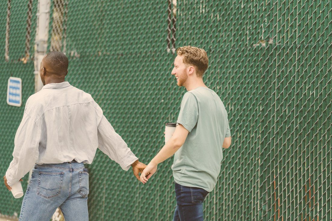 Two men holding hands and smiling on a daytime walk, a warm image of queer connection and affirming care